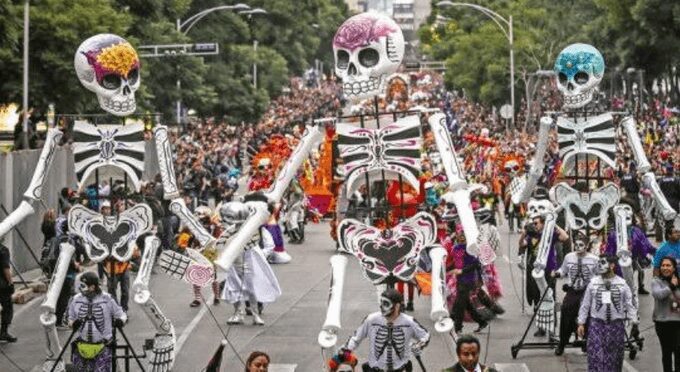Giant colorful skeleton puppets parade down a street filled with spectators.