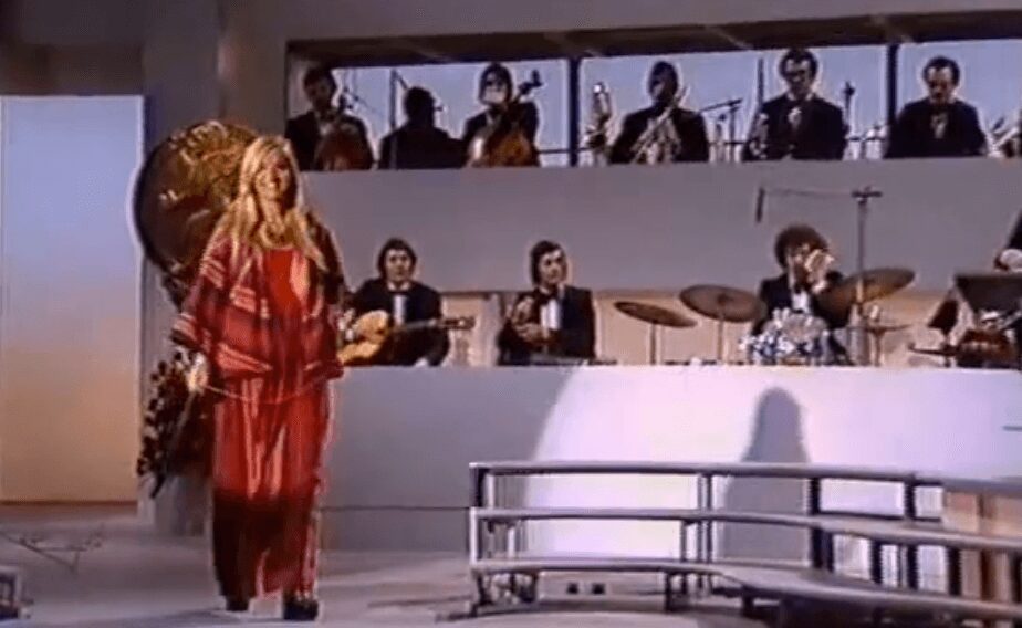 A woman in a red plaid outfit performs on stage with a band behind her.