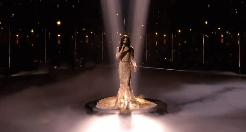 A woman in a shimmering gown sings on a spotlighted stage with fog and city lights.