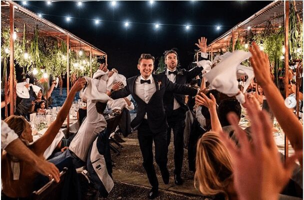 Two men in tuxedos smiling and celebrating at a lively party.