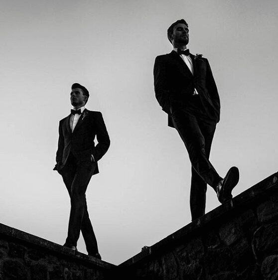 Two men in tuxedos walking confidently on a rooftop at dusk.