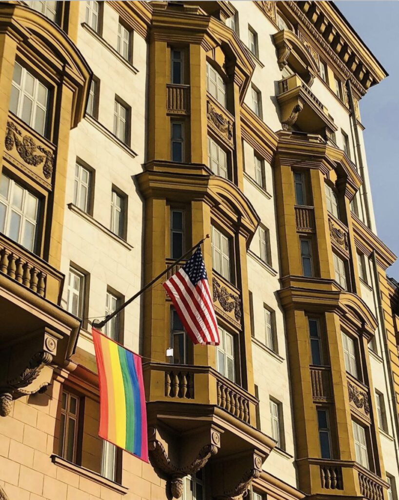 Flags including a rainbow pride and American flag hang from a historic building.